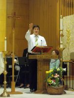 Person in white shirt speaking at a church lectern with raised hand, beside a floral arrangement.