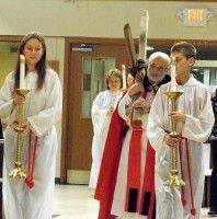 Clergy procession in a church hallway, with candle bearers in white robes and a priest in red vestments.