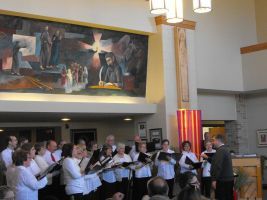 Choir performing in a church-like hall beneath a large mural, with audience seated nearby