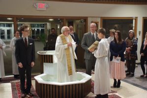 People standing around a lit fountain in a church or hall, with a priest in white vestments at center