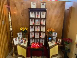 Memorial display with framed photos, flowers, and candles arranged on a table against a wood-paneled wall