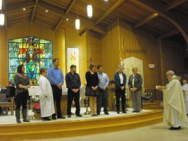 People standing on a church altar during a ceremony, with a priest facing them and stained glass in the background.
