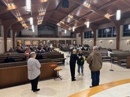Inside a church, a few people stand near pews under a high wooden ceiling.