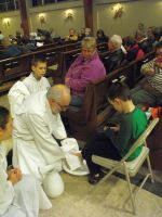 Two clergy kneel beside a seated person in a church, with a congregation seated in pews behind them.