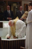 Person in white robes kneels at an altar during a religious ceremony, with others standing nearby.