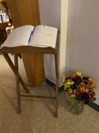 Open book on a wooden stand beside a vase of colorful flowers on a carpeted floor