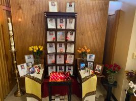 Memorial display with photos, flowers, and candles arranged on a table against a wood-paneled wall.