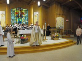Church altar service with clergy in white robes and a choir before a stained-glass window