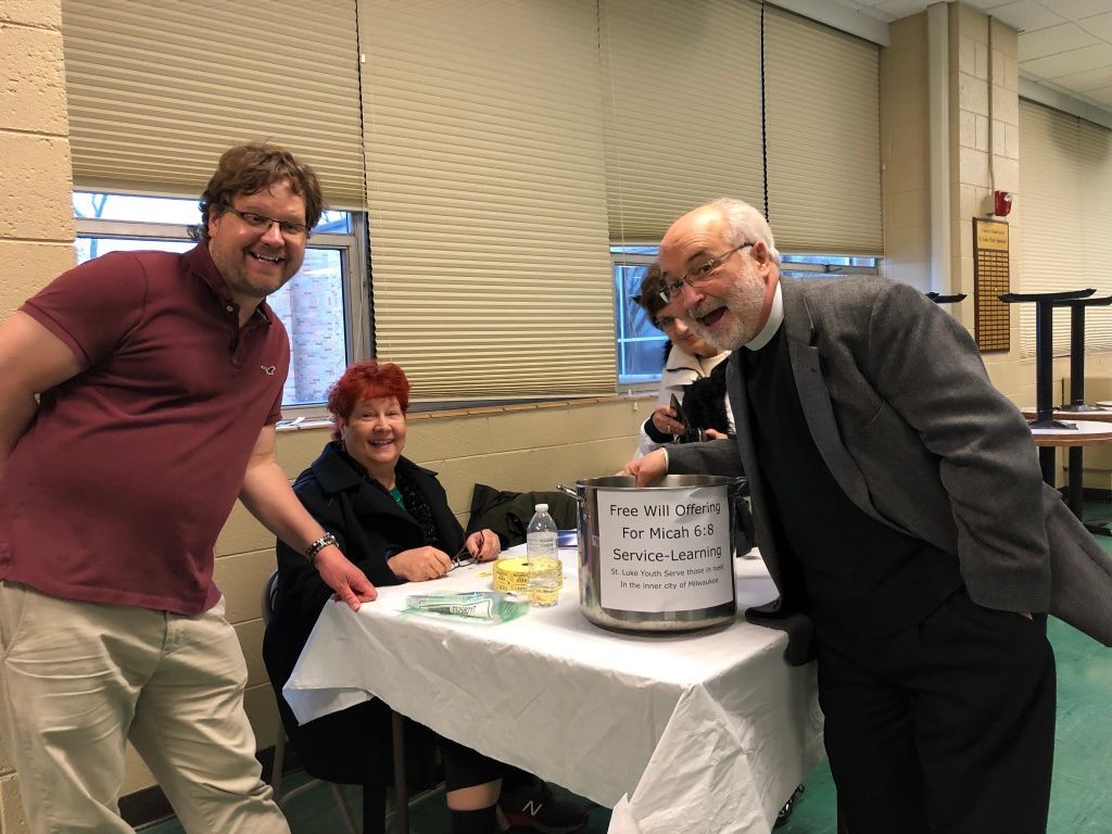 Three people posing beside a donation bucket at a table in an indoor event room.