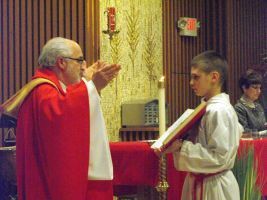 Priest in red robes blessing an altar server in white robes inside a church