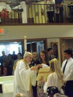 Priest in white robes greeting attendees at a church ceremony, with people gathered around altar candles.