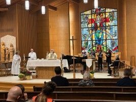 Church service at altar with clergy in white robes, musicians, and a colorful stained-glass window backdrop