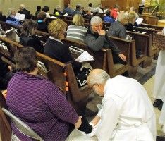 Man kneeling to help a seated person in a church pew, with others gathered around in the background.