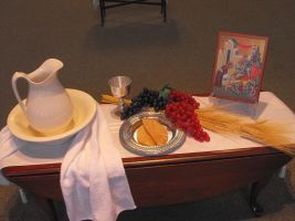 Table setting with white pitcher, cup, fruit, bread, and framed artwork on a cloth-covered table