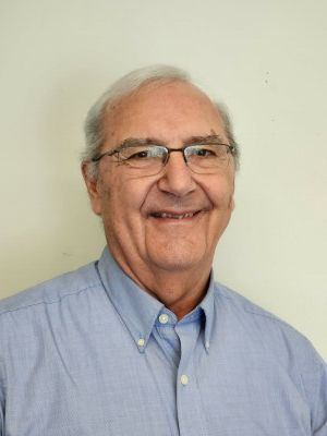 Smiling man in glasses and a light blue button-down shirt against a plain beige background