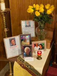 Memorial table with framed photos, yellow flowers, and a lit candle on a cream cloth.