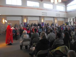 Red-robed speaker addressing a seated crowd in a bright indoor hall