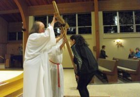 Two people in white robes lifting a wooden cross indoors near pews and seated onlookers.