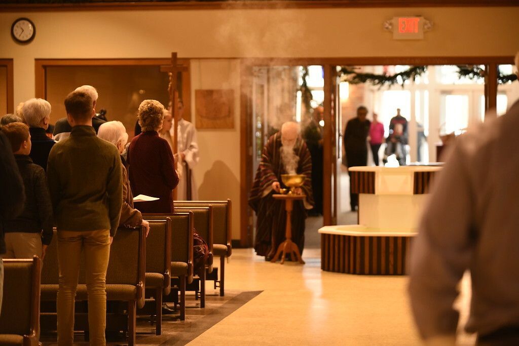 People standing in a church hall during a ceremony, with a bright doorway and altar at the front.