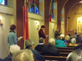 People seated in a church, with a speaker standing near the aisle and stained-glass windows in the background