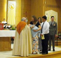 Priest blessing a baby held by a woman during a church baptism ceremony, with family standing nearby.