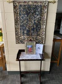 Small memorial table with framed photos and floral cloth beneath a hanging tapestry in a hallway
