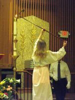 Person in a white dress raises a pole toward a gold panel while a suited man stands behind in a church.
