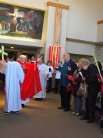 Clergy in red and white robes process through a church aisle beside a standing congregation.