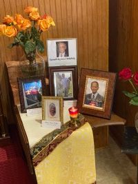 Memorial table with framed photos, flowers, and lit candles against a wood-paneled wall