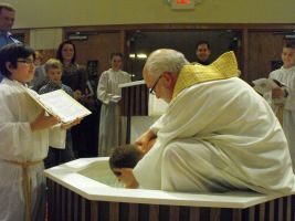 Priest in white robe kneels at altar during church ceremony while readers stand nearby
