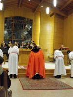 Clergy in robes stand at the altar during a church service, with one in a red cape centered on a rug.