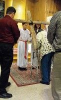 People in a church stand around a priest in white vestments during a blessing ceremony.
