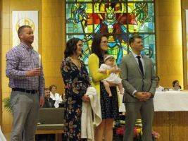 People standing in a church before a stained-glass window, one holding a baby during a ceremony.