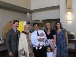 Group posing indoors with a priest, man holding a baby, and two children in front of church walls.
