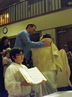 Two people in white robes hold open books during a church ceremony, with a man standing behind them.