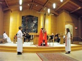 People standing in a church sanctuary during a ceremony, with a red-robed officiant at the altar.