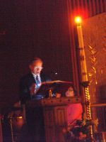 A man stands behind a podium at a lit outdoor night event, with an orange light pole nearby.