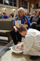 Person kneeling before seated elder, washing feet in a church service with onlookers nearby