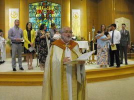 Priest speaking at altar in church, with choir and stained-glass window behind him.
