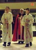 Three people in ceremonial robes and crowns standing on a stage, one wearing a red cape