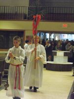 Children in white robes carrying a tall red processional cross indoors