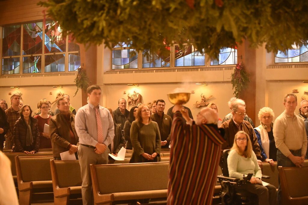 People standing in a church-like hall with pews, warm lighting, and a person in a striped robe in the foreground