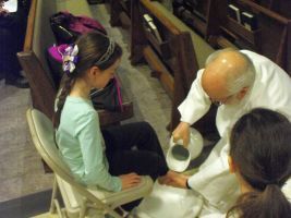 Two people in white clothing pour liquid into a seated child’s foot in a church pew