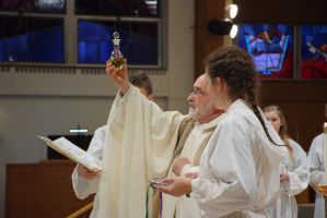 Priest elevates a chalice during a church service as others stand nearby.