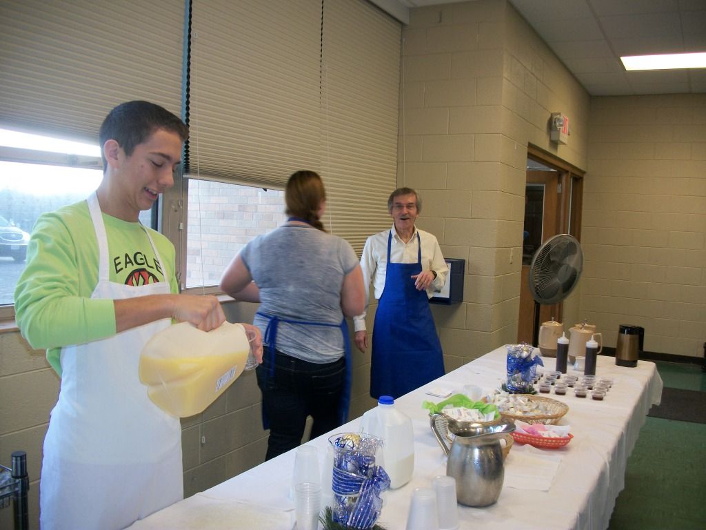 People serving food at a buffet table in a hallway, with bowls, cups, and utensils arranged on the table.