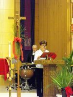 Woman speaking at a church lectern with a drummer in the background, beside a large cross and floral decorations.