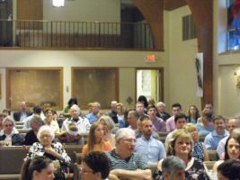 Audience seated in a church or hall, facing forward beneath a balcony.