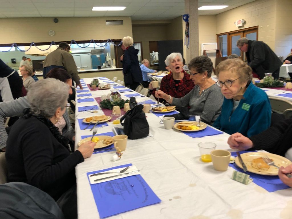 People seated at a long table in a community hall, eating a shared meal with blue napkins and plates.