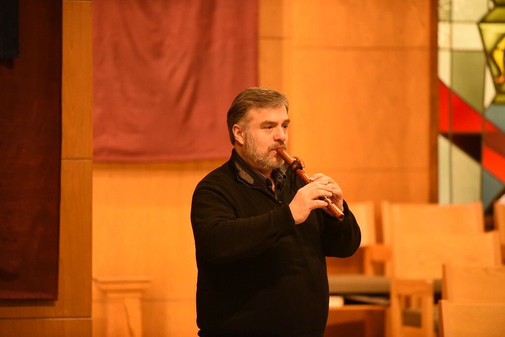 Man speaking with hand gestures in a warmly lit church interior, with wooden pews and stained glass nearby.