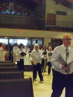 People standing in a church or hall aisle during a gathering, holding books or papers.